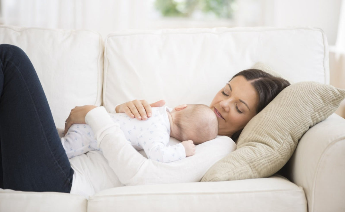 A mother rests on a sofa with her sleeping baby on her chest