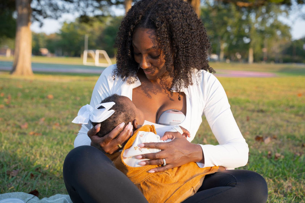 A mother sits on the grass in a park, feeding her baby under a cover