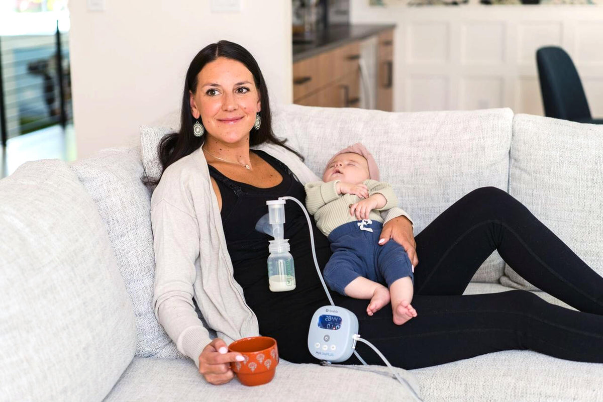 A mother relaxes on a sofa beside her baby while expressing with an Ameda double electric breast pump