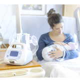 Mother holding her newborn beside the Ameda Pearl breast pump and flanges on a bedside table